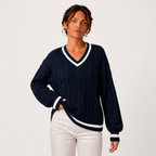 A woman wearing a navy blue cable-knit V-neck sweater with white trim, paired with white pants, photographed against a plain light gray studio background.