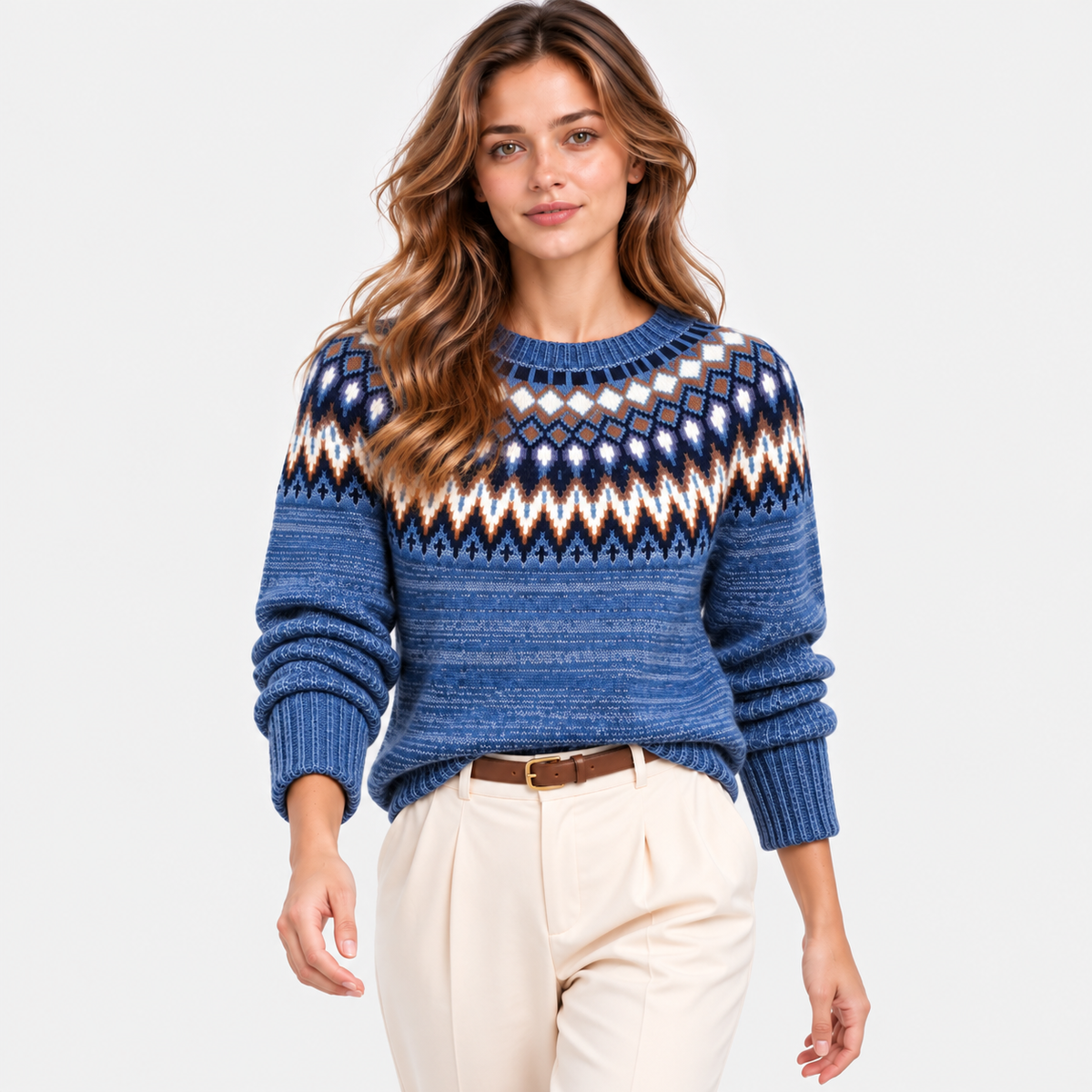 A woman modeling a blue Fair Isle knit sweater with geometric patterns in white, navy, and brown, paired with cream trousers, against a light gray studio background.