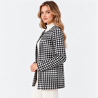 A woman modeling a black and white houndstooth patterned blazer over a white mock-neck top and white pants, photographed against a clean white studio background.