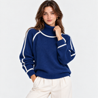 A woman wearing a navy blue knit turtleneck sweater with white contrast stitching along the shoulders, sleeves, and collar, paired with off-white trousers, against a plain light gray background.