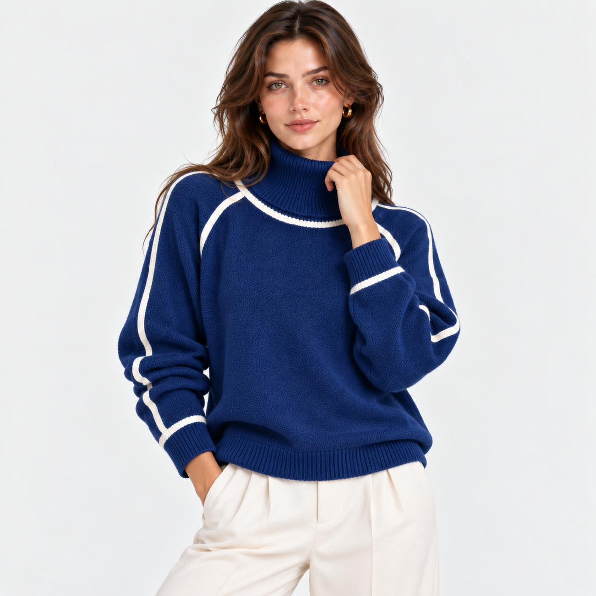 A woman wearing a navy blue knit turtleneck sweater with white contrast stitching along the shoulders, sleeves, and collar, paired with off-white trousers, against a plain light gray background.
