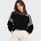 A woman wearing a black knit sweater with white geometric patterns on the shoulders and collar, paired with cream trousers, against a light gray studio background.