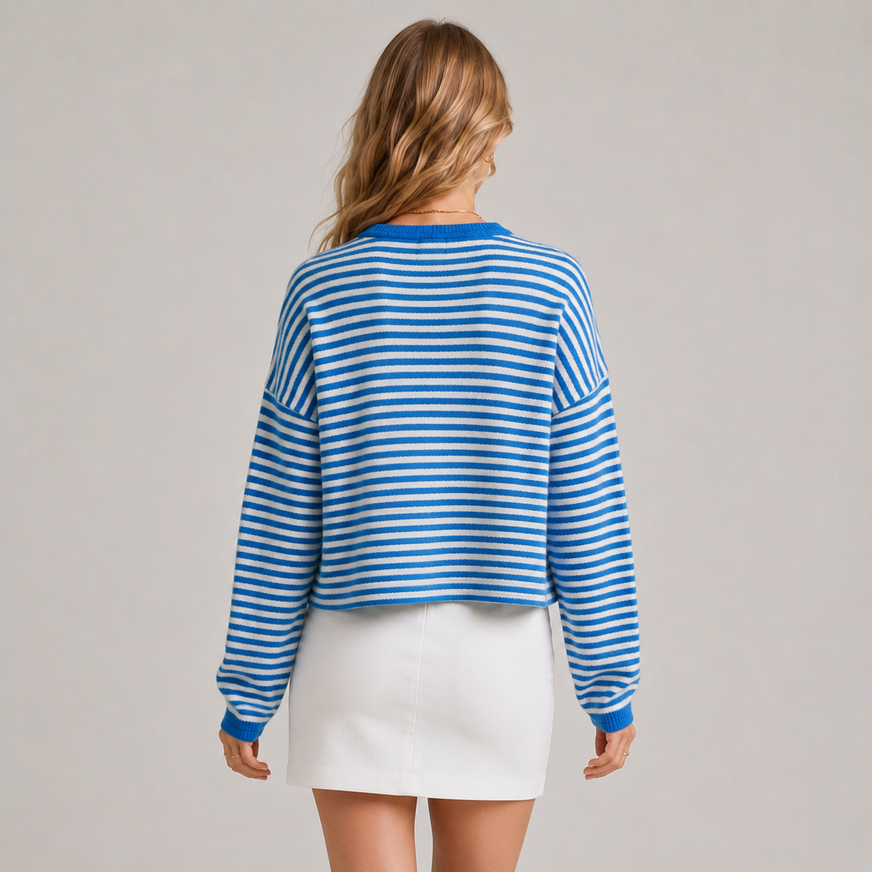 A woman viewed from the back wearing a blue and white horizontally striped long-sleeve crop top and a white mini skirt, set against a plain light gray studio background.