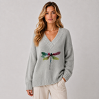 A woman wearing a light gray knit sweater with a colorful dragonfly graphic on the front, paired with cream pants, against a neutral gray background.