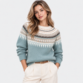 A woman modeling a light blue Fair Isle knit sweater with cream, brown, and beige geometric patterns across the chest and shoulders, paired with cream trousers, against a light gray studio background.