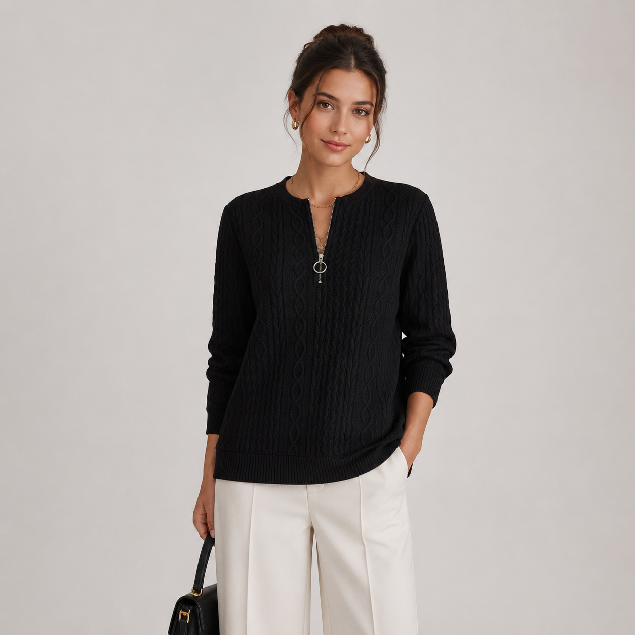 A woman modeling a black ribbed knit sweater with a half-zip neckline, paired with white trousers, against a plain light gray studio background.