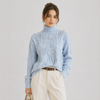 A woman models a light blue cable-knit turtleneck sweater, paired with cream trousers and a brown belt, against a neutral light gray background.