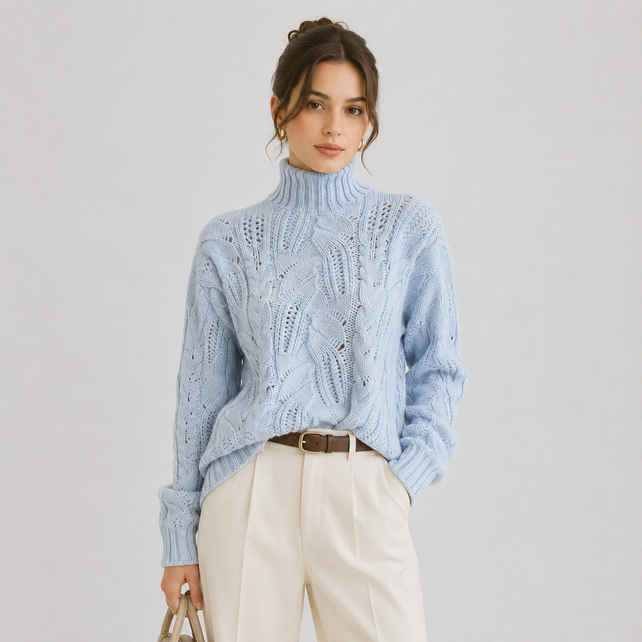 A woman models a light blue cable-knit turtleneck sweater, paired with cream trousers and a brown belt, against a neutral light gray background.