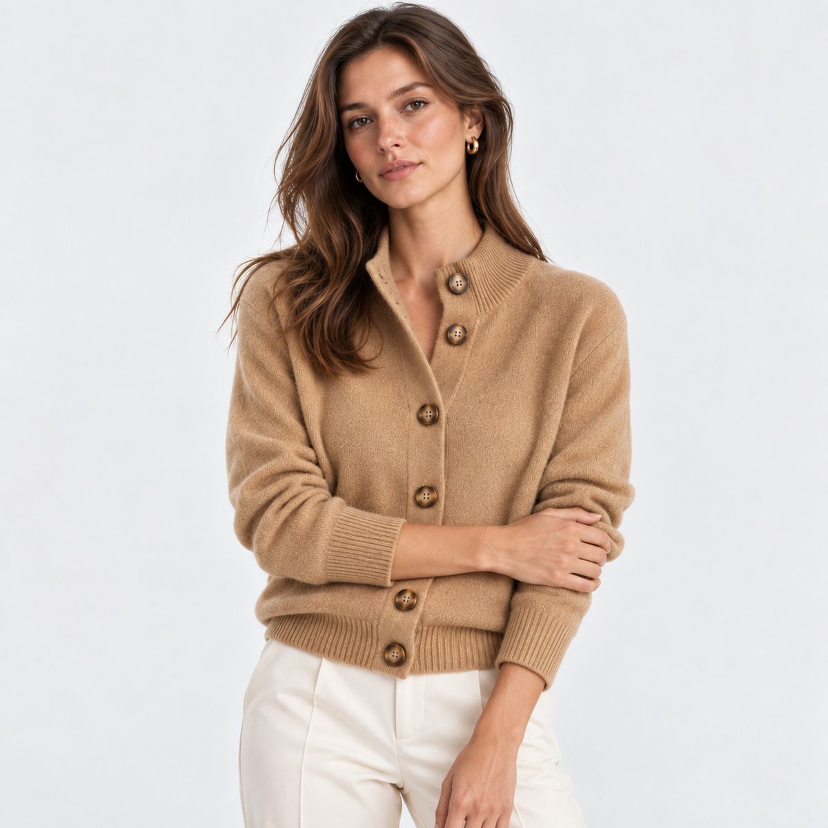 A woman wearing a camel-colored knit cardigan sweater with brown buttons, paired with white pants, photographed against a clean white studio background.