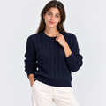 A woman wearing a navy blue cable-knit crewneck sweater and off-white trousers, posing against a light gray studio background.