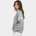 A woman wearing a light gray knit sweater with a white geometric pattern on the upper sleeve, paired with off-white trousers, photographed against a plain light gray studio background.