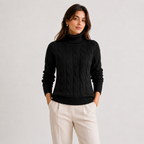 A woman wearing a black cable-knit turtleneck sweater and cream trousers, standing against a light gray studio background.