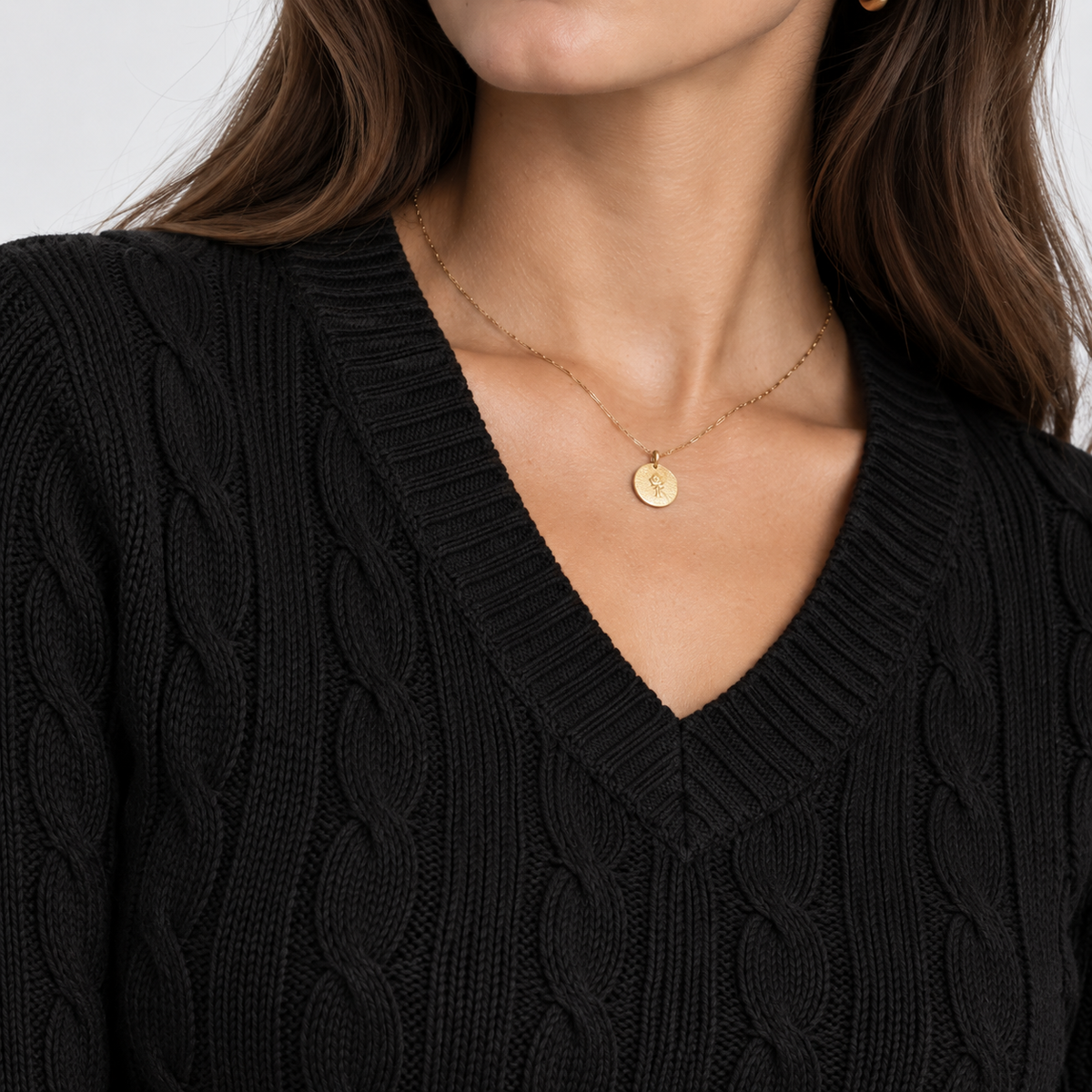 A close-up of a woman wearing a delicate gold chain necklace with a small round disc pendant, styled with a black cable-knit V-neck sweater.