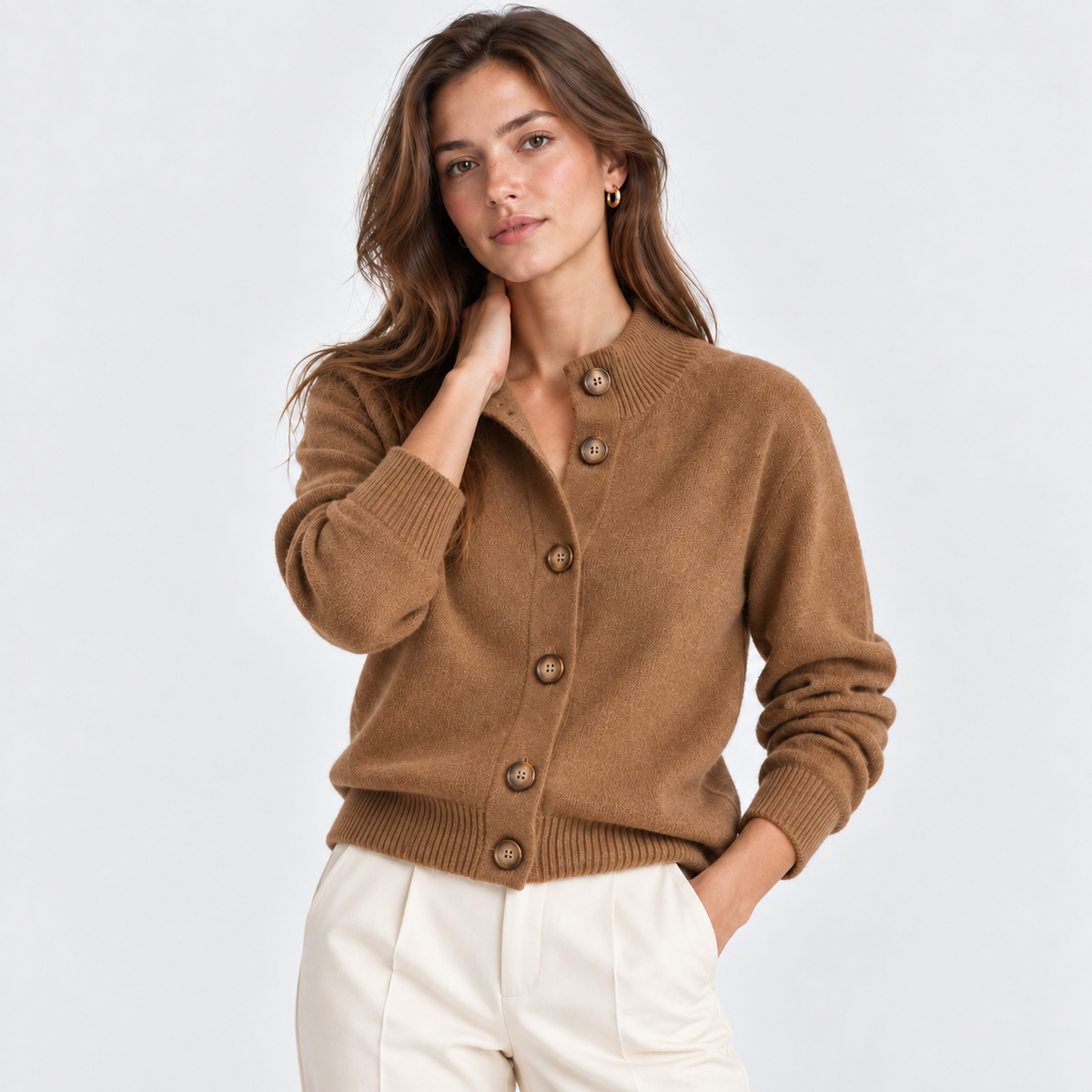 A woman modeling a camel brown button-up cardigan sweater with ribbed collar, cuffs, and hem, paired with off-white trousers, against a light gray studio background.