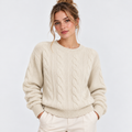 A woman wearing a cream cable-knit crewneck sweater and matching cream trousers, standing against a light gray studio background.