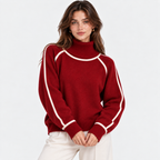 A woman wearing a red mock-neck knit sweater with contrasting white trim along the collar, shoulders, and sleeves, paired with white pants, against a plain white studio background.