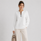 A woman modeling a white textured knit half-zip sweater with a stand collar, paired with light beige wide-leg trousers and holding a taupe handbag, set against a plain light gray studio background.