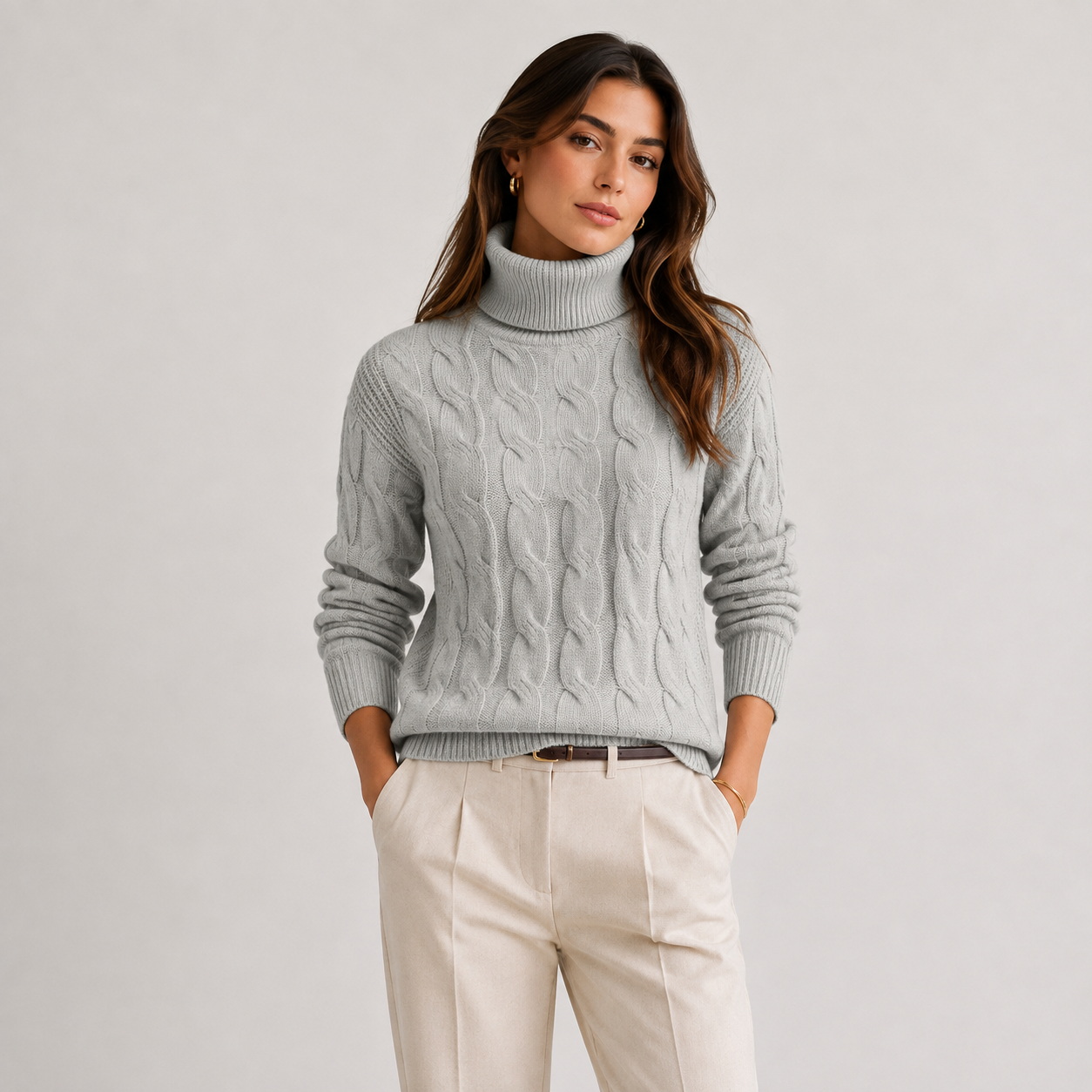 A woman wearing a light gray cable-knit turtleneck sweater and cream-colored trousers, standing against a neutral light gray studio background.