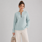 A woman models a light mint green cable-knit sweater with a half-zip neckline, paired with cream wide-leg trousers and holding a beige handbag, set against a plain light gray studio background.