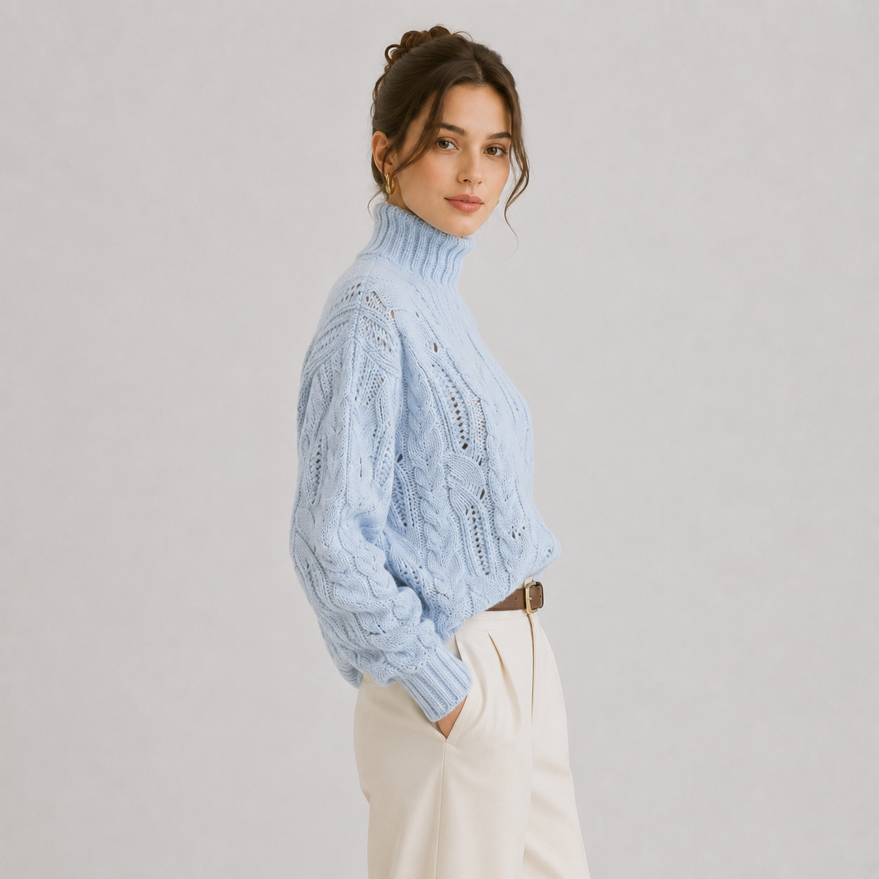 A woman wearing a light blue cable-knit turtleneck sweater and cream trousers, photographed against a plain light gray studio background.
