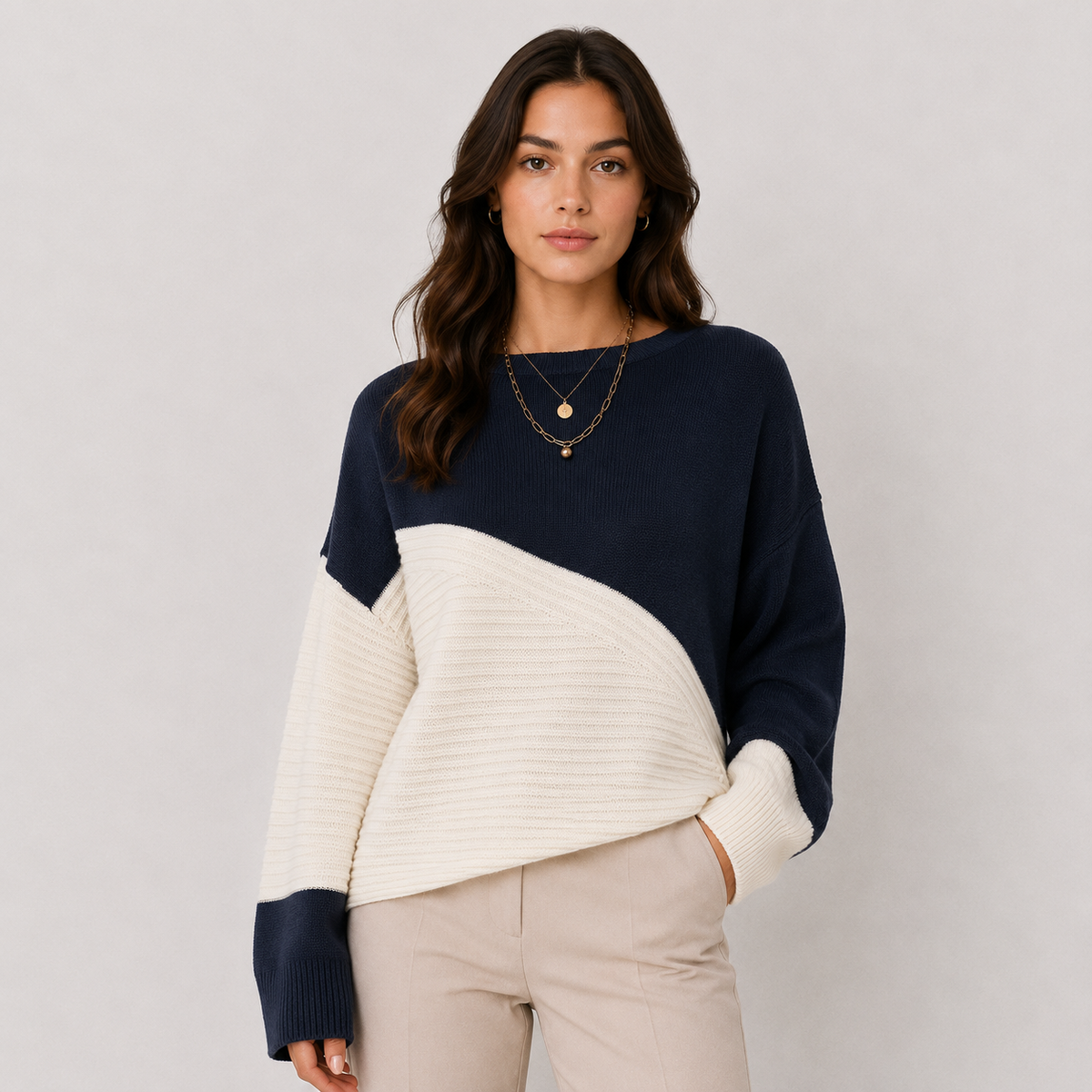 A woman modeling a color-block knit sweater with a navy blue upper section and sleeves, and a cream ribbed lower section and sleeves, paired with light beige pants against a light gray studio background.