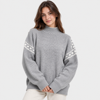 A woman wearing a light gray knit sweater with white geometric patterns on the sleeves, paired with off-white pants, against a plain light gray background.
