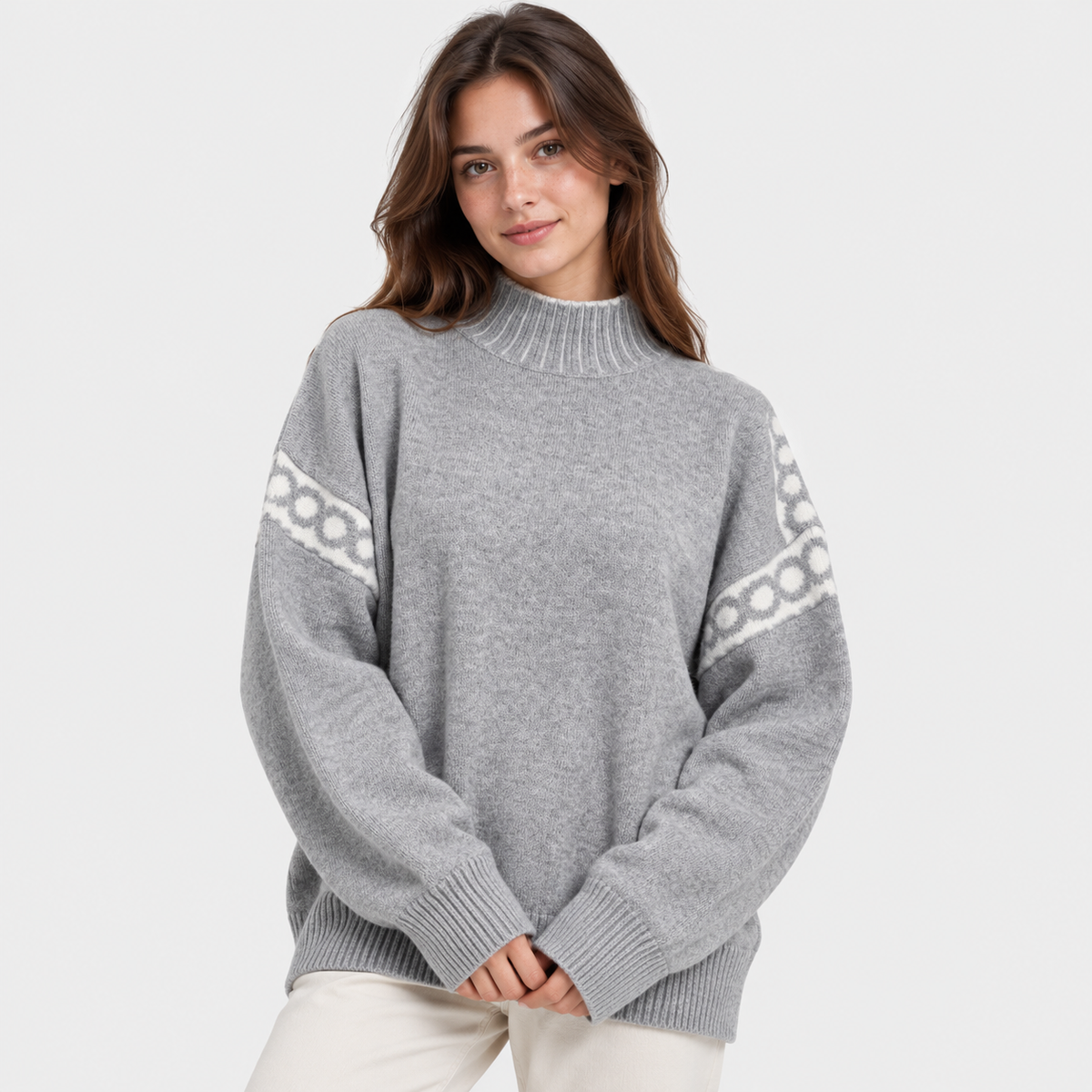 A woman wearing a light gray knit sweater with white geometric patterns on the sleeves, paired with off-white pants, against a plain light gray background.