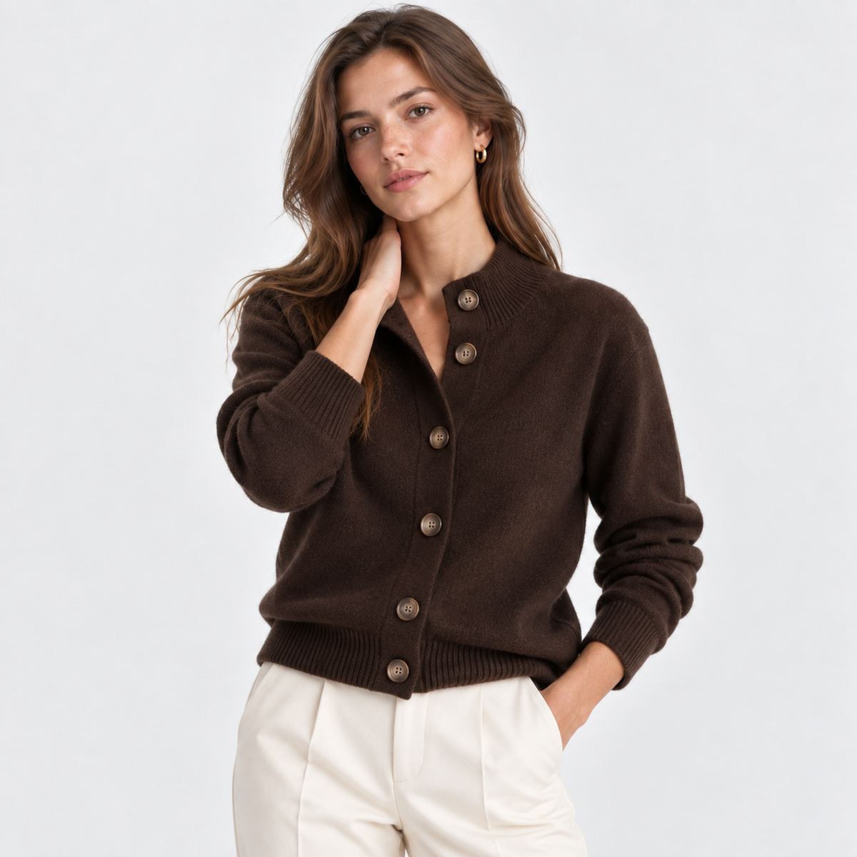 A woman modeling a dark brown knit cardigan sweater with brown buttons, paired with cream-colored pants, against a plain light gray studio background.