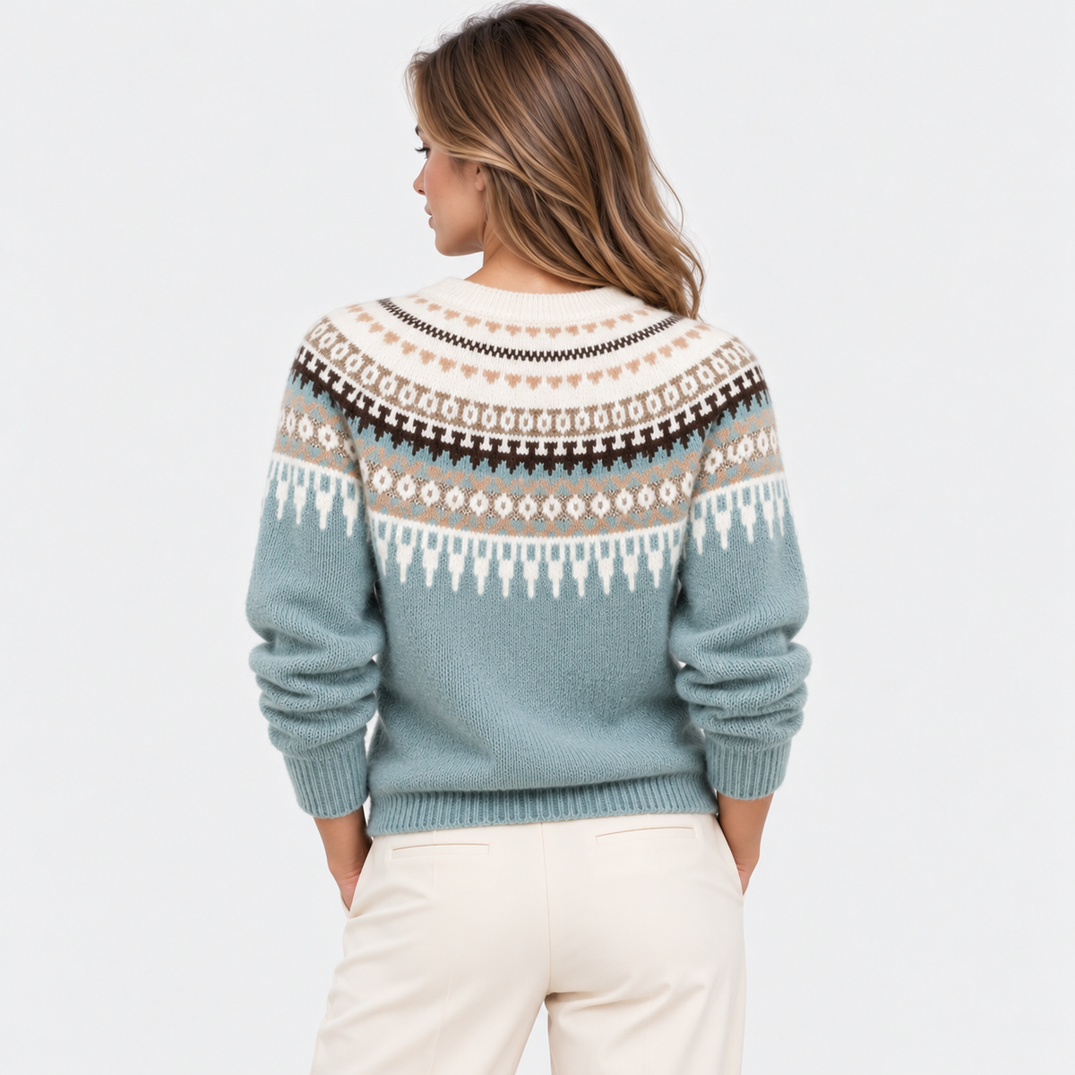 A woman viewed from the back wearing a knit sweater with a multicolor Fair Isle pattern in sage green, cream, brown, and light blue, paired with cream pants, against a light gray studio background.