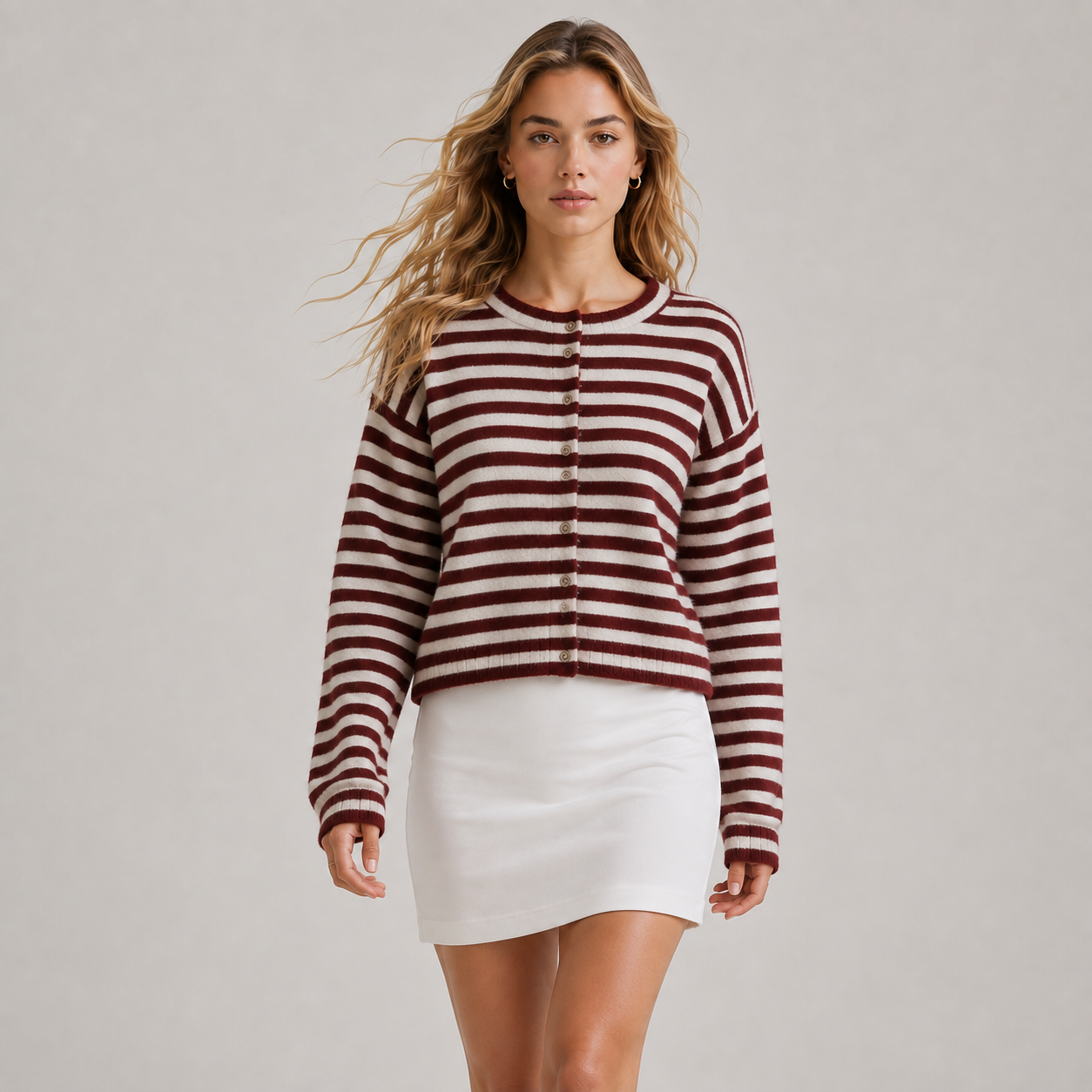 A woman modeling a cropped, long-sleeve knit sweater with horizontal burgundy and off-white stripes, featuring a button-up front, paired with a white mini skirt, against a neutral light gray background.