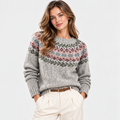 A woman wearing a gray knit crewneck sweater with a multicolor Fair Isle floral yoke pattern, paired with cream trousers and a brown belt, against a plain light gray studio background.