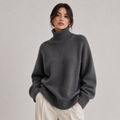 A woman wearing an oversized dark gray ribbed turtleneck sweater and cream-colored trousers, standing against a neutral light gray studio background.