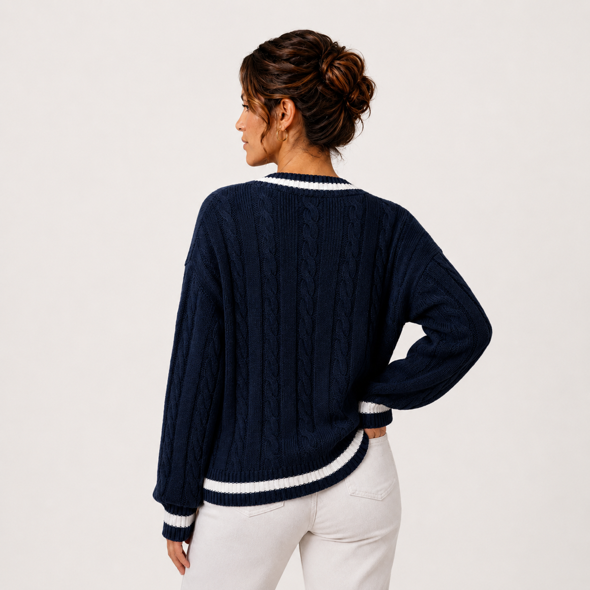 A woman shown from the back wearing a navy blue cable-knit sweater with white ribbed trim at the collar, cuffs, and hem, paired with white pants, against a plain off-white studio background.