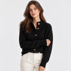 A woman modeling a black knit cardigan sweater with brown buttons, paired with cream trousers, against a light gray studio background.