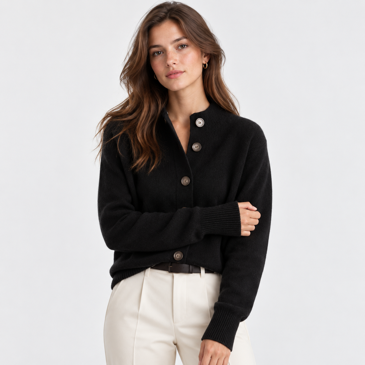 A woman modeling a black knit cardigan sweater with brown buttons, paired with cream trousers, against a light gray studio background.
