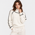 A woman wearing a cream-colored knit mock-neck sweater with black contrast piping along the collar, sleeve cuffs, and shoulders, paired with matching cream sweatpants, set against a plain light gray background.