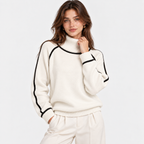 A woman wearing a cream-colored knit mock-neck sweater with black contrast piping along the collar, sleeve cuffs, and shoulders, paired with matching cream sweatpants, set against a plain light gray background.