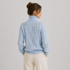 A woman shown from the back wearing a light blue cable-knit turtleneck sweater and cream-colored pants, set against a plain light gray studio background.