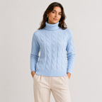 A woman wearing a light blue cable-knit turtleneck sweater and cream trousers, standing against a plain light gray studio background.