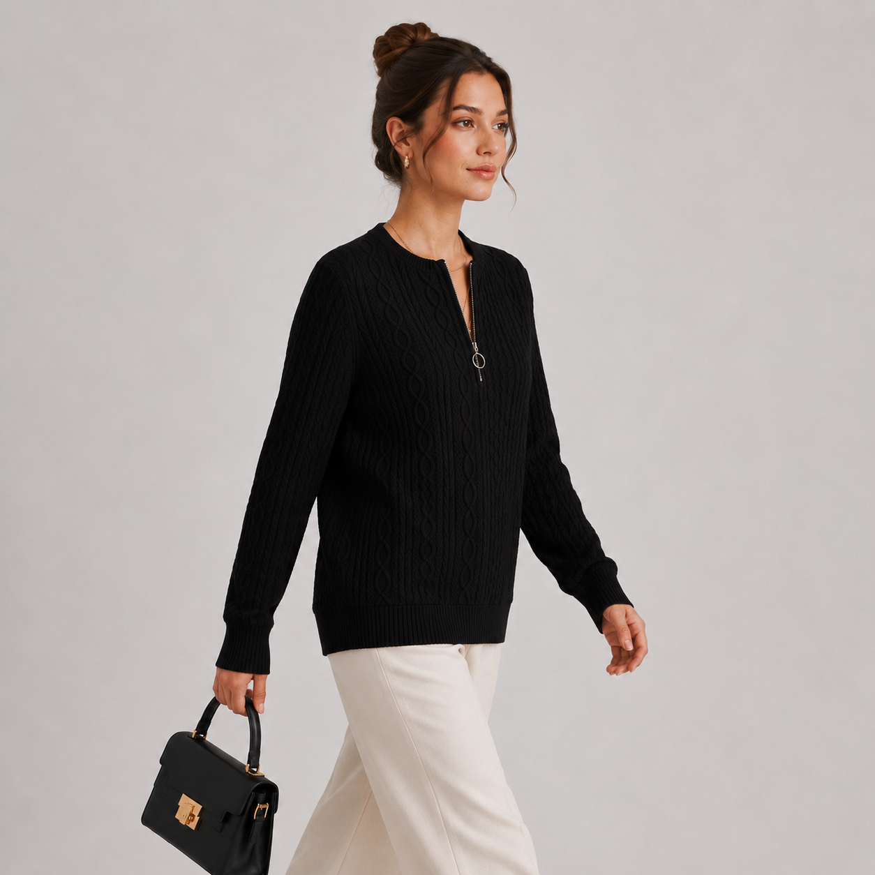 A woman wearing a black ribbed knit sweater with a split V-neckline and zipper detail, paired with white trousers and carrying a black handbag, set against a plain light gray studio background.