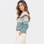 A woman wearing a light blue knit sweater with a cream and black Fair Isle pattern across the yoke and sleeves, paired with cream trousers, against a plain white studio background.