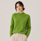 A woman wearing a bright lime green knit mock neck sweater and light beige trousers, standing against a solid light gray background.