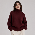 A woman wearing an oversized ribbed turtleneck sweater in deep burgundy, paired with cream-colored trousers, photographed against a neutral light gray studio background.