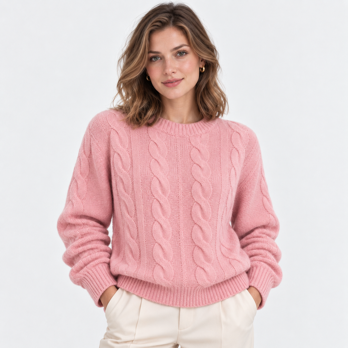 A woman wearing a pink cable-knit crewneck sweater and cream-colored trousers, standing against a plain white studio background.