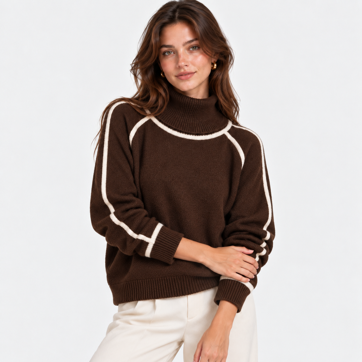 A woman wearing a brown mock neck knit sweater with contrasting cream-colored piping details on the shoulders and sleeves, paired with cream-colored pants, set against a plain light gray background.