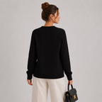 A woman shown from the back wearing a black cable-knit long-sleeve sweater and cream-colored trousers, holding a small black handbag against a light gray studio background.