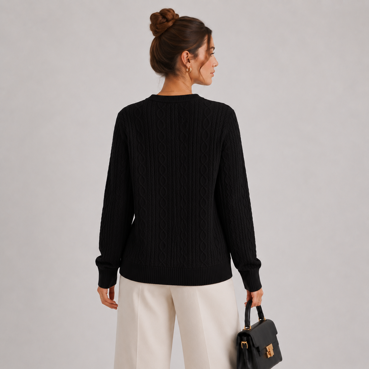 A woman shown from the back wearing a black cable-knit long-sleeve sweater and cream-colored trousers, holding a small black handbag against a light gray studio background.