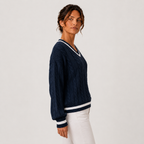 A woman wearing a navy blue cable-knit sweater with white striped trim on the collar, cuffs, and hem, paired with white pants, photographed against a plain light gray studio background.