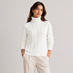 A woman wearing a white cable-knit turtleneck sweater and light beige trousers, standing against a plain light gray studio background.