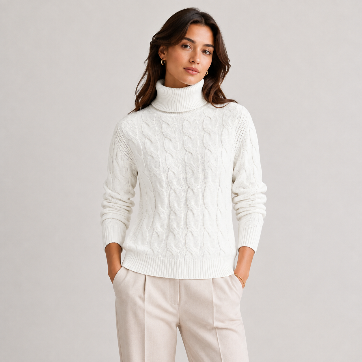 A woman wearing a white cable-knit turtleneck sweater and light beige trousers, standing against a plain light gray studio background.